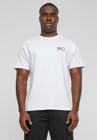 White cotton t-shirt with short sleeves, featuring a small black graphic on the left chest. Model wearing black cargo pants, standing upright.