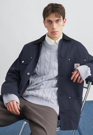 Fiatal férfi szemüvegben, réteges kötött pulóverben, sötét dzsekiben és barna nadrágban, laza testtartással ül egy székben, egyszínű háttér előtt.