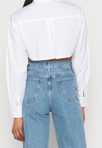 Camisa blanca recortada de botones con mangas largas, que presenta puños con abertura y un detalle en la parte posterior, combinada con unos jeans de mezclilla azul de tiro alto.