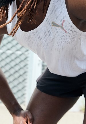 Nahaufnahme einer Person mit Dreadlocks, die ein weißes Puma-Sporttanktop und schwarze Shorts trägt und sich im Freien nach vorne beugt.