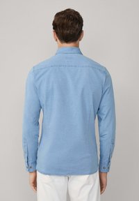 Hombre de espaldas vistiendo una camisa de botones de manga larga azul claro y pantalones blancos frente a un fondo simple.