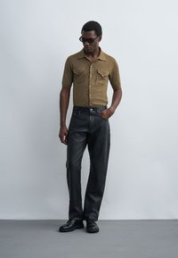 Homme portant une chemise marron à manches courtes boutonnée, un pantalon en cuir noir taille haute, des chaussures noires et des lunettes de soleil noires, debout devant un fond uni.