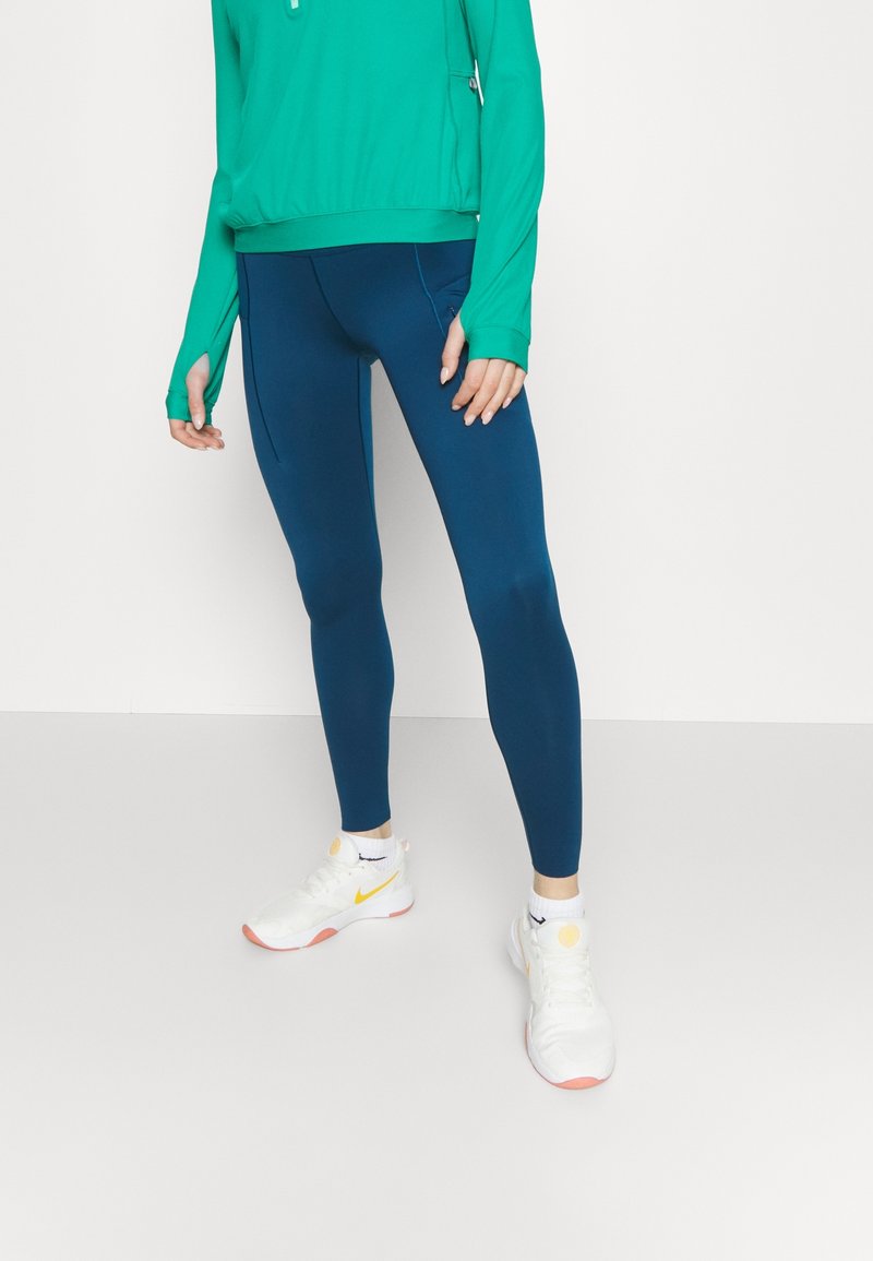 Top verde a maniche lunghe con aperture per i pollici, abbinato a leggings blu scuro. Scarpe da ginnastica bianche con accenti gialli e rosa. Tessuto morbido.