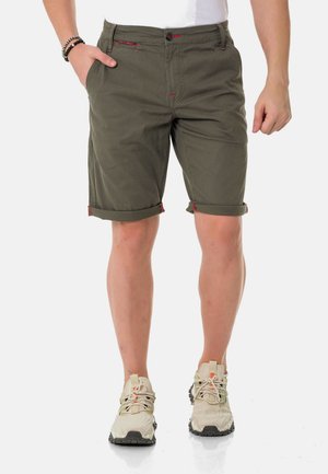 Olivgrüne Baumwollshorts mit umgeschlagenen Säumen, ausgestattet mit kontrastierendem roten Nähten und einem Knopfverschluss. Getragen mit hellen Sport Schuhen.