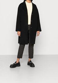 Czarny wełniany płaszcz z klapowanym kołnierzem, o luźnym kroju i z trzema guzikami. Połączony z jasnżółtą bluzką, wzorzystymi spodniami i masywnymi czarnymi butami.