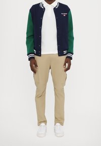 Marinblå och grön varsityjacka med randiga muddar, bärs över en vit polo t-shirt, tillsammans med beige cargobyxor och vita sneakers.