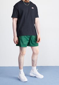 Svart bomullströja med en liten röd logotyp, tillsammans med gröna shorts. Vita sneakers och ankelsockor, stående på ett ljust blått golv.