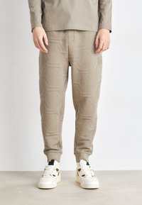 Calças de jogger em bege claro com um padrão geométrico em relevo, punhos elásticos e bolsos laterais, combinadas com ténis brancos com detalhes coloridos.