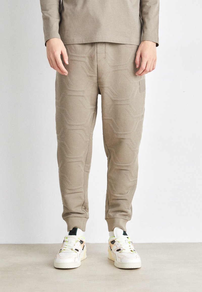 Calças de jogger em bege claro com um padrão geométrico em relevo, punhos elásticos e bolsos laterais, combinadas com ténis brancos com detalhes coloridos.