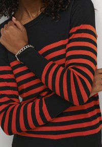 Personne portant un pull rayé noir et rouge horizontalement, les bras croisés et un bracelet chaîne argenté à un poignet.