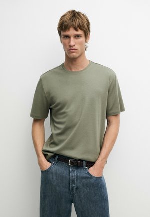 Junger Mann mit hellbraunen Haaren, der ein einfaches grünes kurzärmeliges T-Shirt und blaue Jeans trägt, mit den Händen in den Taschen vor weißem Hintergrund.