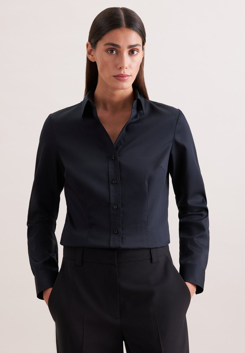 Femme aux cheveux noirs et raides portant une chemise noire cintrée à boutons et un pantalon noir, debout les mains dans les poches devant un fond uni.