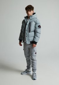 Veste doudoune gris clair avec poches zippées, accents noirs et poignets côtelés, associée à un pantalon de survêtement cargo gris comportant des poches latérales.
