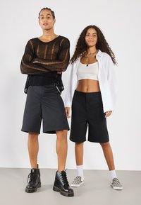 Shorts noirs bien taillés, haut en maille semi-transparente à manches longues noir, débardeur blanc court et chemise blanche oversized. Bottes noires et chaussures à carreaux.