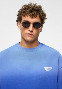Blauwe gradient sweatshirt met ronde hals, met een klein wit logo-tekst. Model draagt zwarte ovale zonnebril en heeft krullend haar.