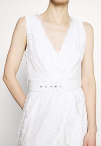 Robe en dentelle blanche avec un décolleté en V et des bretelles larges, dotée d'une ceinture assortie avec une boucle carrée et des éléments en métal argenté. Motif floral texturé.