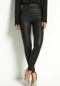 Persona vestita con pantaloni di pelle nera attillati e scarpe con tacchi alti argentati, in piedi con le gambe incrociate e le mani rilassate ai lati.