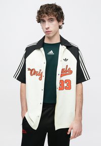 Λευκό και μαύρο πουκάμισο με κουμπιά, με πορτοκαλί κείμενο και τον αριθμό 33, συνδυασμένο με μια σκούρη πράσινη μπλούζα Adidas και μαύρο παντελόνι, με λεπτομέρειες επωνυμίας.