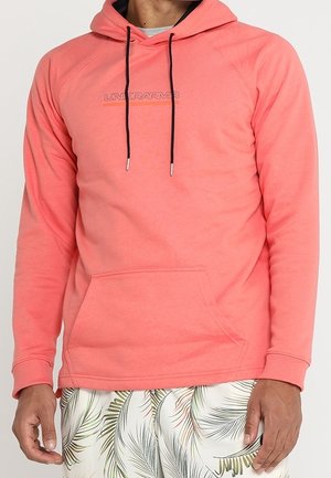 Mężczyzna w koralowo-różowej bluzie z kapturem Under Armour z czarnymi sznurkami oraz białych szortach z zielonym nadrukiem liści palmowych.