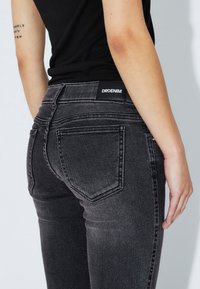 Dark gray denim jeans with a high waist, featuring back pockets and a rectangular black label with "DRDENIM." Smooth texture, slim fit.