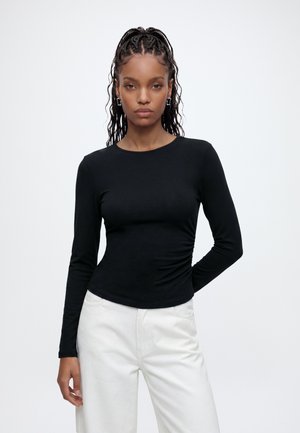 Femme aux cheveux tressés portant des boucles d'oreilles en argent en forme de créoles, un chemisier noir ajusté à manches longues et un pantalon blanc, debout avec une main dans le dos.