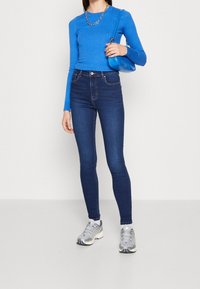 Blau geripptes Langarmoberteil kombiniert mit hoch taillierten, dunklen Jeans im Skinny-Fit. Das Model trägt silberne Sportschuhe und hält eine blaue Handtasche.