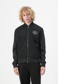 Black bomber jacket with gray sleeves, zip closure, ribbed hem and cuffs, featuring a textured patch on the left chest. Smooth fabric finish.