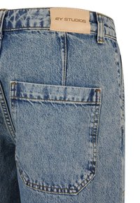 Jean en denim bleu clair montrant la poche arrière et un patch en cuir beige avec le logo "2Y STUDIOS" au-dessus de la poche.