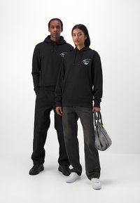Sudaderas negras con bolsillos frontales y acentos gráficos en contraste, combinadas con jeans negros y grises. Un bolso gris con un patrón texturizado.