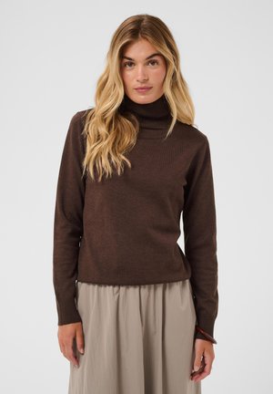 Jeune femme aux longs cheveux blonds portant un pull à col roulé marron foncé et une jupe beige clair sur un fond clair uni.