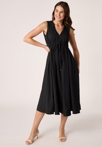Zwarte mouwloze maxi-jurk met V-hals, geplooid middel en strikdetail; gemêleerde stof, vloeiende silhouet en lengte tot onder de knie.