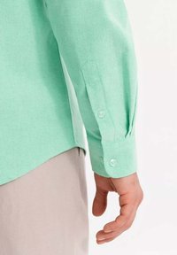 Camicia button-up verde chiaro con maniche lunghe, dotata di un polsino con due bottoni e un tessuto liscio e strutturato. Mostrata su una mano.