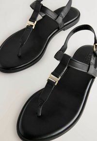 Sandalias planas negras con diseño de tira entre los dedos, confeccionadas en cuero suave, con tiras finas y un acento de hardware en tono dorado.