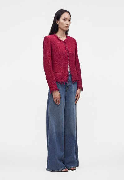 Femme debout dans une veste rouge texturée à bords effilochés, pantalon en jean bleu à jambes larges et sandales noires, sur un fond uni clair.