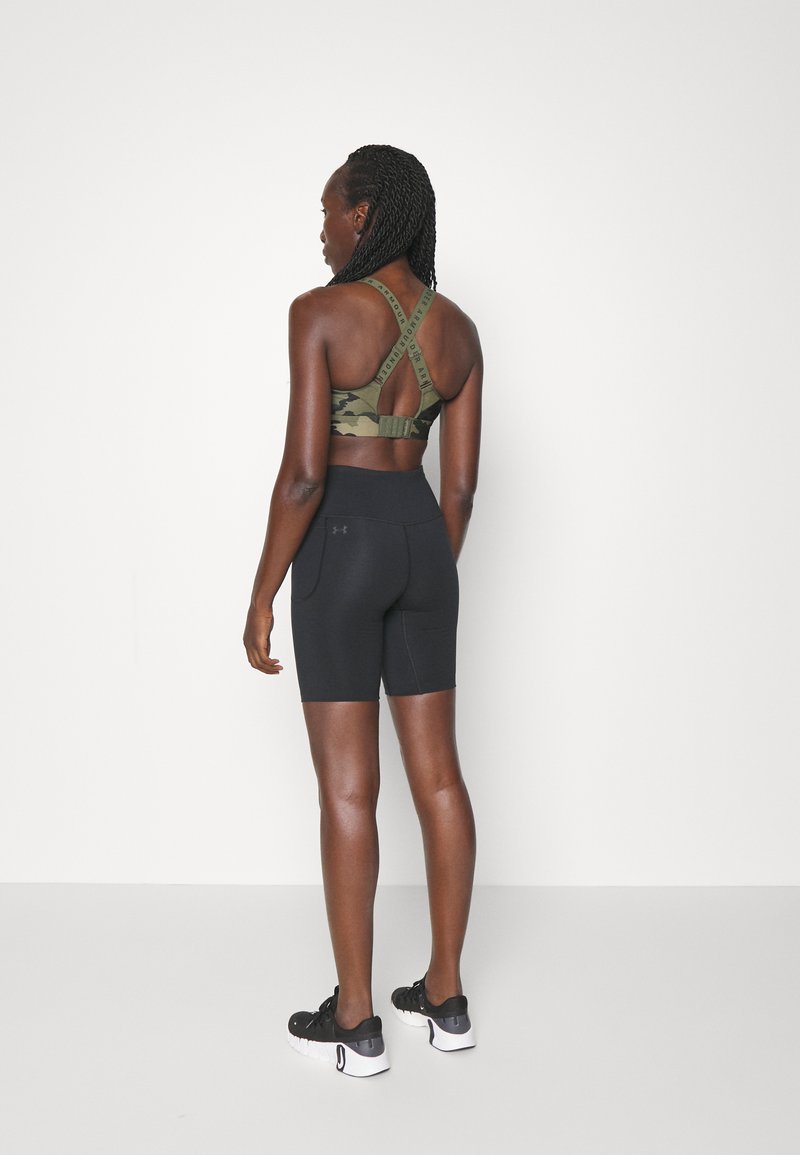 Mujer con trenzas que lleva un sujetador deportivo de camuflaje verde y pantalones cortos atléticos negros de cintura alta, de pie frente a una pared blanca.