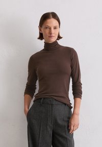 Frau mit kurzem braunem Haar, die einen braunen Rollkragenpullover und graue Hochbundhosen trägt, steht vor einer schlichten hellen Wand.
