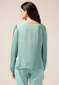 Blusa a maniche lunghe di colore verde menta in tessuto leggero, con un ampio scollo e morbidi drappeggi sulle spalle, dotata di un orlo curvo.