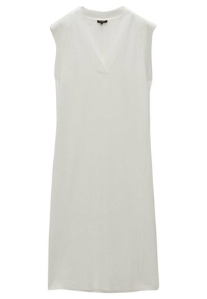Woman wearing a sleeveless white midi dress with a deep V-neckline and black flat sandals, standing against a plain white wall.