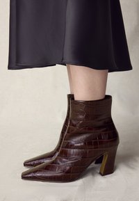 Braune Stiefeletten aus krokodilledergeprägtem Leder mit spitzer Schuhspitze und Blockabsatz. Sie zeichnen sich durch eine strukturierte Oberfläche und eine schlanke Silhouette aus.