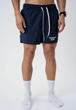 Man wearing navy blue shorts with white drawstrings and white text, white socks, and a black wristwatch on a plain white background.