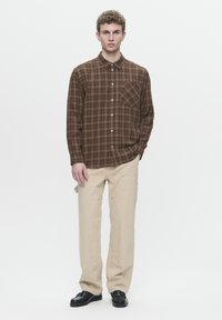 Chemise à manches longues à carreaux marron avec fermeture à boutons et poche. Associée à un pantalon beige et des chaussures noires. Design simple et structuré.