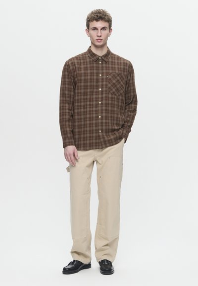 Chemise à manches longues à carreaux marron avec fermeture à boutons et poche. Associée à un pantalon beige et des chaussures noires. Design simple et structuré.