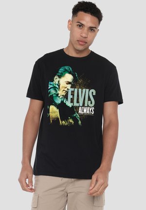 Young man wearing a black t-shirt with a colored graphic of Elvis Presley playing guitar and text "ELVIS ALWAYS The Original."