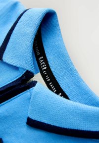 Golier svetlomodrého polokošele s tmavomodrým lemovaním a viditeľnou značkou "BAKER by TED BAKER" vo vnútri goliera.