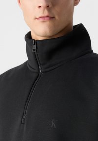 Felpa nera con mezza zip caratterizzata da un colletto alto, texture morbida e discreto logo CK sul lato sinistro del petto. Design semplice e moderno.