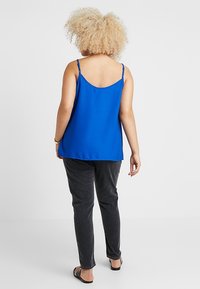 Modrý bezrukávový top s volným střihem a zaobleným výstřihem, kombinovaný s tmavě šedými kalhotami. Hedvábná látka s nastavitelnými ramínky.