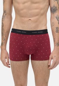 Torso masculino vistiendo bóxer rojos con pequeñas huellas de patas blancas y una cinturilla negra etiquetada "Ysabel Mora," mostrando tatuajes en los brazos.