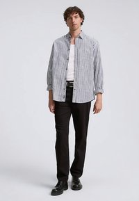 Črtasta srajca s knofki v beli in mornarsko modri barvi, udobnega kroja z zavihanimi rokavi; kombinirana s črnimi hlačami in vezalkami obutimi čevlji.