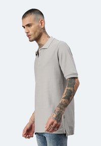 Polo shirt grigio a maniche corte realizzato in tessuto testurizzato, dotato di colletto con bottoni e spacchi laterali, indossato da un modello con tatuaggi.