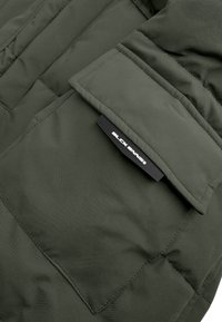 Olijfgroene gewatteerde jas met een rechthoekige zak, uitgerust met een zwart label met "BLCK BNDS" in witte tekst, met een gladde textuur en stikdetails.